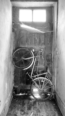 A dilapidated bicycle is leaning up against a wooden door inside an old, dusty room. The bicycle is missing several parts and is surrounded by cobwebs and debris. Light filters through a small window at the top of the door, creating a stark contrast between the bright window and the shadowy interior.
