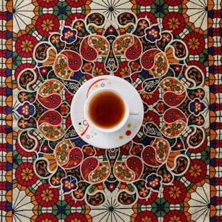 A white teacup filled with tea sits on an intricately patterned tablecloth. The design features ornate floral and paisley motifs in vibrant colors including red, orange, green, and blue, creating a rich and detailed background.