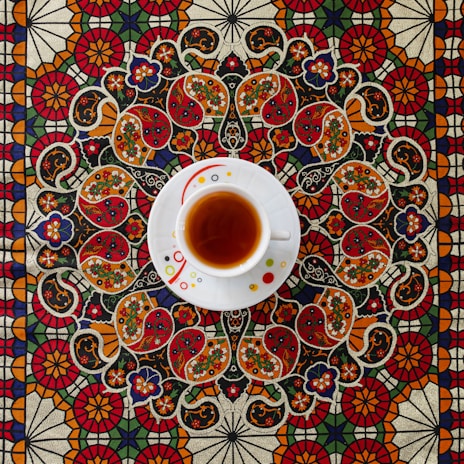 A white teacup filled with tea sits on an intricately patterned tablecloth. The design features ornate floral and paisley motifs in vibrant colors including red, orange, green, and blue, creating a rich and detailed background.