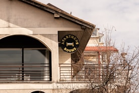 A sign for apartments and rooms is mounted on the exterior of a building. The structure has a large arch-shaped window with metal railings. Leafless branches of a tree are visible in the foreground. The building features a slanted roof and multiple levels.