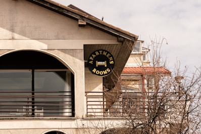 A sign for apartments and rooms is mounted on the exterior of a building. The structure has a large arch-shaped window with metal railings. Leafless branches of a tree are visible in the foreground. The building features a slanted roof and multiple levels.