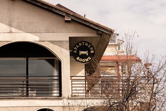 A sign for apartments and rooms is mounted on the exterior of a building. The structure has a large arch-shaped window with metal railings. Leafless branches of a tree are visible in the foreground. The building features a slanted roof and multiple levels.