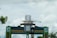 A large, decorative gate features a model ship on top, surrounded by vibrant green panels with inscriptions. Palm trees and a cloudy sky form the backdrop.
