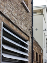 A narrow alleyway between two buildings showcasing a brick facade. A security camera is mounted on a black pipe attached to the brick wall. The windows have white louvered shutters angled inwards, allowing light while maintaining some privacy. The background shows a modern building with smooth, light-colored walls.
