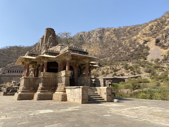 A historical stone temple structure with intricate carvings is set in a mountainous landscape. The temple features a series of columns and a stepped entrance, with a prominent tower rising above it. Surrounding the temple is a vast stone courtyard, and the background displays a dry, rocky hillside dotted with sparse vegetation under a clear blue sky.