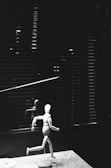 A wooden artist mannequin is positioned in a running pose on a flat surface. Behind the mannequin, there is a window covered with horizontal blinds, casting subtle shadows. The scene is captured in black and white, giving it a dramatic and abstract feel.