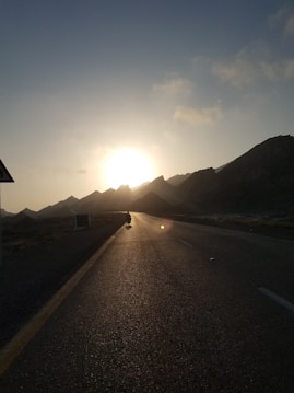 A motorcyclist rides along a road with a dramatic sunset in the background. The silhouette of mountains is visible under the clear sky, with soft clouds adding to the scenery. The sunlight reflects off the road, casting a warm glow.