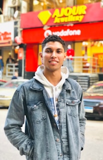 A young person wearing a denim jacket over a hoodie stands in a busy urban street. Behind them are storefronts with bright red and yellow signage and a few pedestrians. Vehicles are parked along the street.
