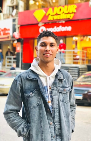 A young person wearing a denim jacket over a hoodie stands in a busy urban street. Behind them are storefronts with bright red and yellow signage and a few pedestrians. Vehicles are parked along the street.
