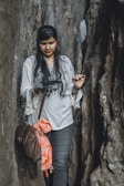 A woman with long, dark hair is standing between two large rock formations, holding a camera gimbal with a smartphone attached. She is wearing a white top with intricate embroidery and jeans. A brown bag is slung over her shoulder, and an orange patterned cloth is tied to it.