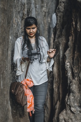 A woman with long, dark hair is standing between two large rock formations, holding a camera gimbal with a smartphone attached. She is wearing a white top with intricate embroidery and jeans. A brown bag is slung over her shoulder, and an orange patterned cloth is tied to it.