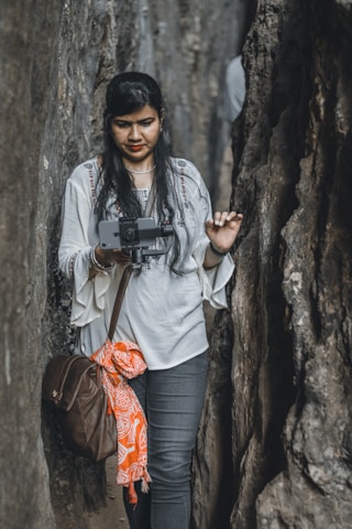 A woman with long, dark hair is standing between two large rock formations, holding a camera gimbal with a smartphone attached. She is wearing a white top with intricate embroidery and jeans. A brown bag is slung over her shoulder, and an orange patterned cloth is tied to it.