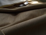A close-up view of a textured fabric with a beige or light brown color, draped softly with shadows and light creating gentle contours. The weave pattern is clearly visible, showcasing the intricate detail of the material. In the background, there is a hint of a shiny or reflective object, partially obscured, adding an element of interest.