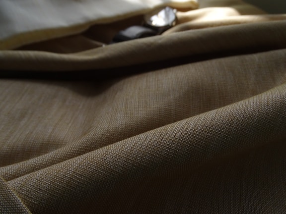 A close-up view of a textured fabric with a beige or light brown color, draped softly with shadows and light creating gentle contours. The weave pattern is clearly visible, showcasing the intricate detail of the material. In the background, there is a hint of a shiny or reflective object, partially obscured, adding an element of interest.