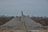 A large mound of gravel is present, with a conveyor belt structure extending over it. The background features a line of bare trees beneath a cloudy sky. The surrounding area appears industrial and earthy, with utility poles and sparse vegetation.