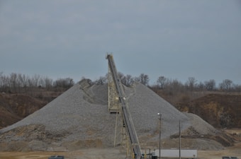 A large mound of gravel is present, with a conveyor belt structure extending over it. The background features a line of bare trees beneath a cloudy sky. The surrounding area appears industrial and earthy, with utility poles and sparse vegetation.