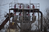 A metal structure with pipes and machinery rises above leafless branches. The structure includes several large cylindrical components, and there are platforms and railings surrounding it. The sky is overcast, adding a somber atmosphere.