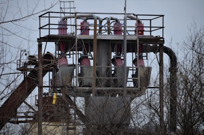A metal structure with pipes and machinery rises above leafless branches. The structure includes several large cylindrical components, and there are platforms and railings surrounding it. The sky is overcast, adding a somber atmosphere.