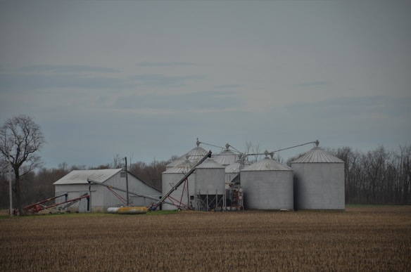 Several large metal grain silos and a warehouse building are situated in an agricultural field. The silos are equipped with extended conveyors, likely for loading and unloading grains. The field appears to be plowed or ready for planting, and bare trees are visible in the background under a cloudy sky.