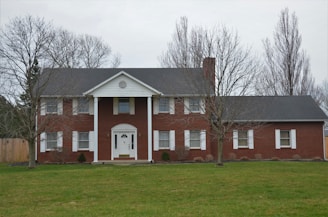 A two-story red brick house with white columns and trim is situated in a grassy yard. The roof is dark gray, and the house features a central doorway with a semi-circular fanlight and sidelights. There are several windows with white shutters, and leafless trees frame the scene.