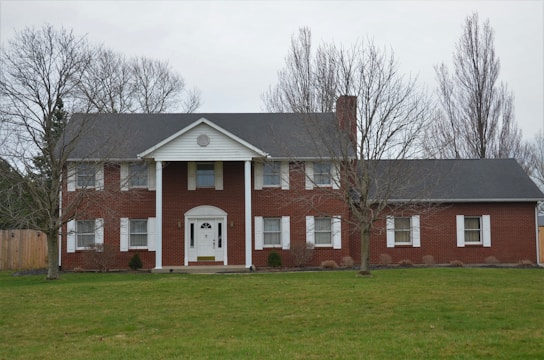 A two-story red brick house with white columns and trim is situated in a grassy yard. The roof is dark gray, and the house features a central doorway with a semi-circular fanlight and sidelights. There are several windows with white shutters, and leafless trees frame the scene.
