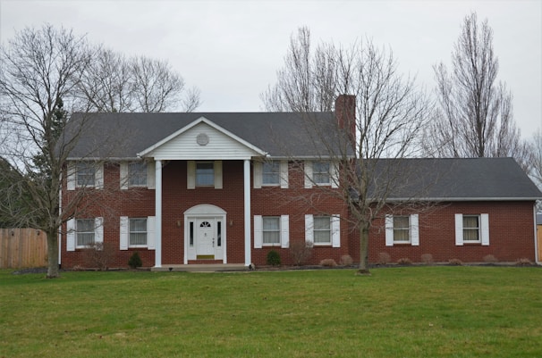 A two-story red brick house with white columns and trim is situated in a grassy yard. The roof is dark gray, and the house features a central doorway with a semi-circular fanlight and sidelights. There are several windows with white shutters, and leafless trees frame the scene.