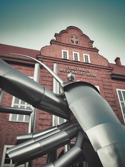 A large modern metallic sculpture is positioned in front of a historical brick building. The sculpture features abstract, angular shapes with a futuristic aesthetic. The building is adorned with architectural details from an earlier period and has an inscription above the entrance.