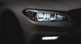 A close-up view of a car's front end focusing on its modern, sleek headlight with bright LED lights shining in a dimly lit setting. The car's body is mostly in shadow, emphasizing the sharp design and reflective elements of the headlight.