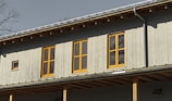 A building facade with vertical wood paneling, featuring several yellow-framed windows reflecting sunlight. The roof has visible rafters and a metal gutter, and bare tree branches can be seen in the background.