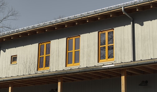 A building facade with vertical wood paneling, featuring several yellow-framed windows reflecting sunlight. The roof has visible rafters and a metal gutter, and bare tree branches can be seen in the background.