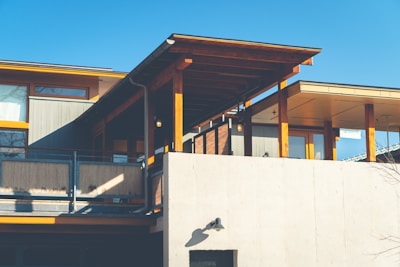 A modern house with a combination of wood and concrete materials. It features large windows, wooden beams, and a concrete exterior wall. The overhanging roof provides shade and adds architectural interest. The sky is clear and blue, indicating a sunny day.