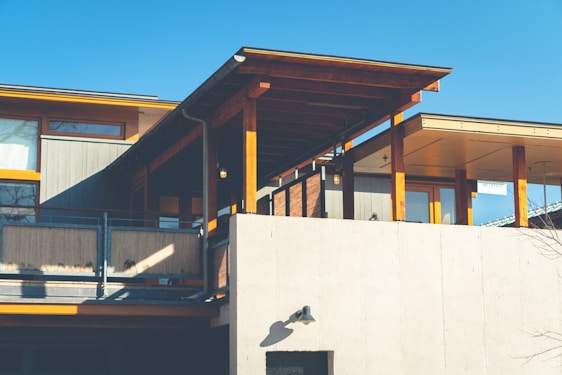 A modern house with a combination of wood and concrete materials. It features large windows, wooden beams, and a concrete exterior wall. The overhanging roof provides shade and adds architectural interest. The sky is clear and blue, indicating a sunny day.