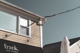 A corner of a building with beige siding and a black awning displaying the text 'frank. food company' is visible. A window with white frames is present above the awning. Overhead wires and a drainpipe are attached to the building's corner. The sky is clear, and part of a white object, possibly an umbrella or structure, is in the foreground.