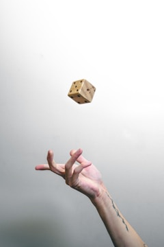 A tattooed hand is playfully tossing a wooden dice in the air against a plain white background.