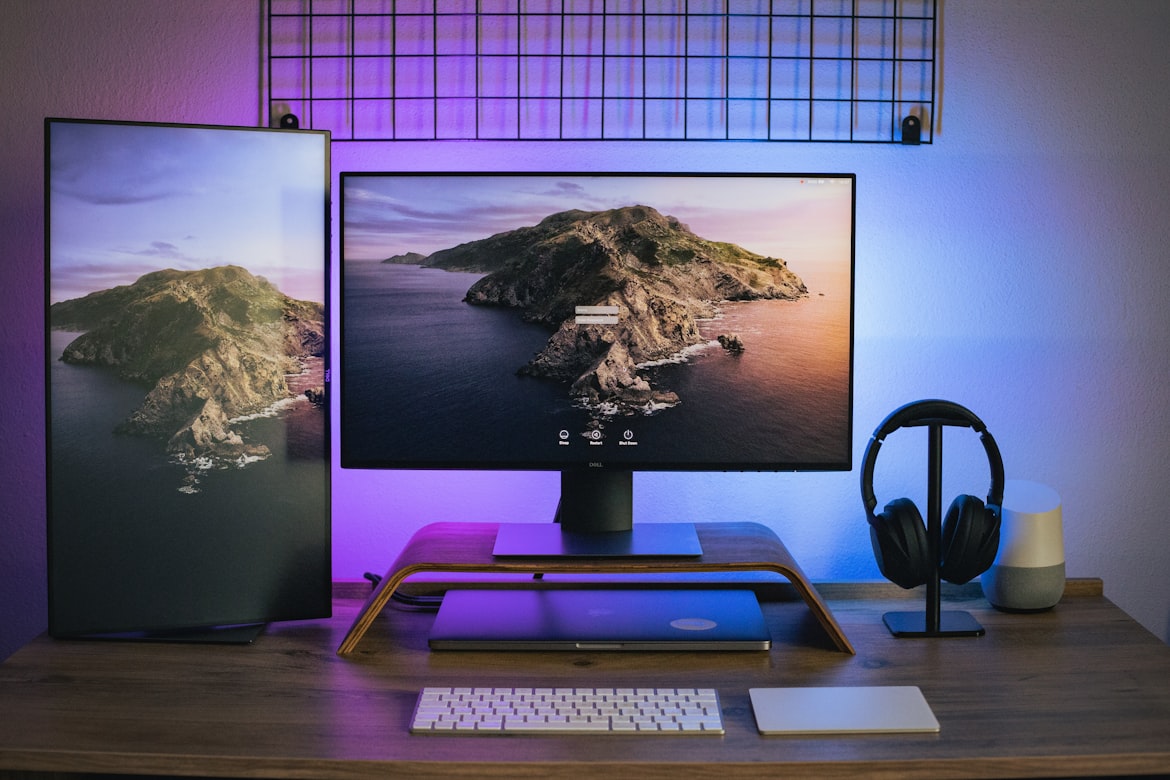 computer with two screens displaying an island on the screensaver, one vertical and other horizontal standing on a wood support, and a headphone all on a table