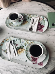 On a marble table, two floral-patterned trays each hold a cup of black coffee, a spoon, and a napkin with the text 'Beymen'. A green smartphone lies nearby, and part of an arm is visible at the top of the image.