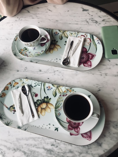 On a marble table, two floral-patterned trays each hold a cup of black coffee, a spoon, and a napkin with the text 'Beymen'. A green smartphone lies nearby, and part of an arm is visible at the top of the image.