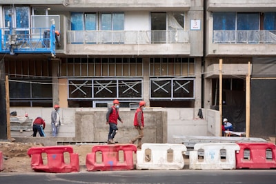 Several construction workers wearing safety gear, including helmets and vests, are engaged in various tasks at a building site. The structure appears to be in the middle stages of construction, with large concrete barriers and scaffolding visible. The building’s windows are partially installed and covered for protection.