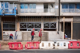 Several construction workers wearing safety gear, including helmets and vests, are engaged in various tasks at a building site. The structure appears to be in the middle stages of construction, with large concrete barriers and scaffolding visible. The building’s windows are partially installed and covered for protection.