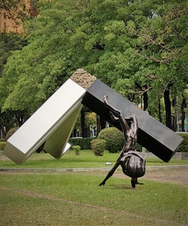 A large outdoor sculpture featuring a metallic human figure performing a handstand, integrated into a geometric structure composed of angular shapes in black and white. Surrounding the sculpture is a grassy area with neatly trimmed bushes and lush green trees, creating a park-like setting.