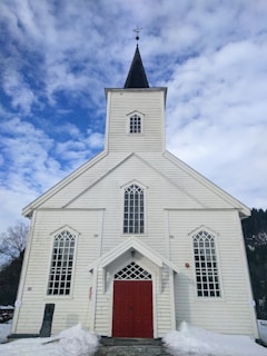 A traditional white wooden church with red double doors and a dark steeple topped with a weather vane. The structure features tall, narrow windows with intricate framing. Snow is present on the ground, and a clear sky above has scattered clouds.
