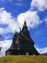A traditional wooden stave church with intricate detailing on its steeply sloped roof, standing on a grassy hill under a bright blue sky filled with scattered white clouds.