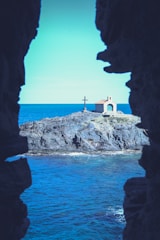 A small chapel with a red roof sits on a rocky outcrop surrounded by the ocean. A large cross stands next to the chapel. The scene is framed by dark rock formations.