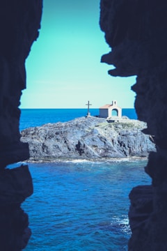 A small chapel with a red roof sits on a rocky outcrop surrounded by the ocean. A large cross stands next to the chapel. The scene is framed by dark rock formations.
