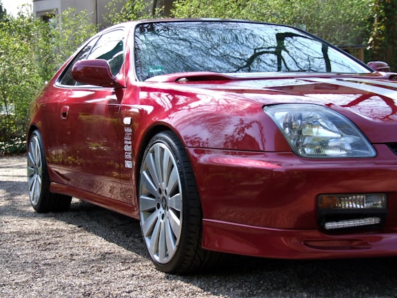 A red sports coupe with sleek, aerodynamic features is parked on a gravel driveway. The car's glossy finish reflects the surrounding greenery and trees. Large alloy wheels and clear headlights emphasize its modern design. The vehicle appears to be well-maintained, with detailed stickers along the side adding a personal touch.