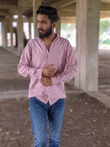 A person with a beard and short hair is standing in a partially constructed building. They are wearing a light pink buttoned shirt and blue jeans. The area around them is open and unfinished, with concrete pillars and a dirt floor.
