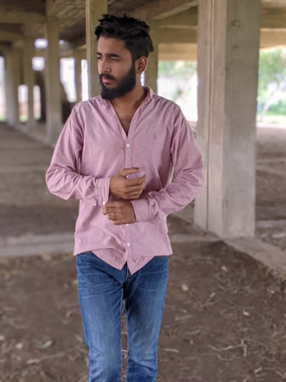 A person with a beard and short hair is standing in a partially constructed building. They are wearing a light pink buttoned shirt and blue jeans. The area around them is open and unfinished, with concrete pillars and a dirt floor.