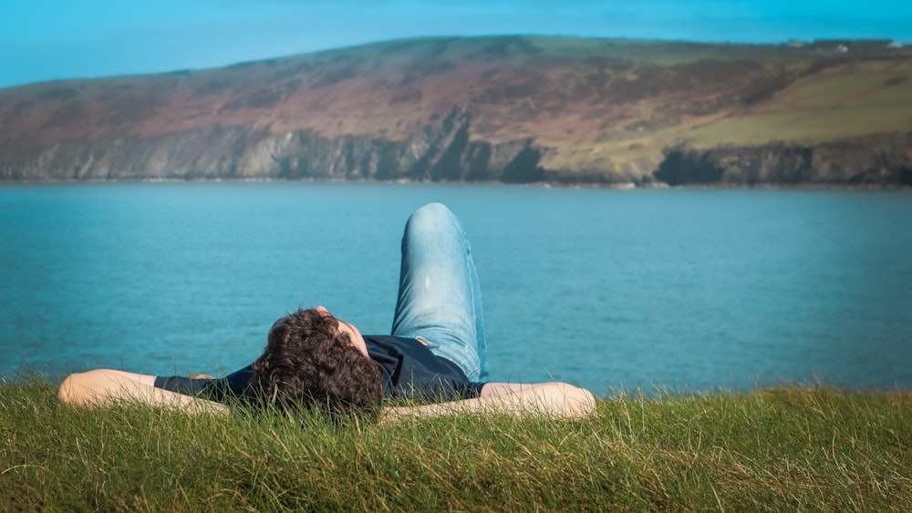 Persona con camisa negra y jeans de mezclilla azules sentada en una roca marrón cerca del cuerpo de agua