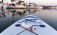 A stand-up paddleboard rests on calm waters in a marina, with several boats docked in the background. The paddleboard has a logo on it, and a cylindrical object with a purple ring is placed on it, secured by a rope. The sun is setting, casting a serene light over the scene.