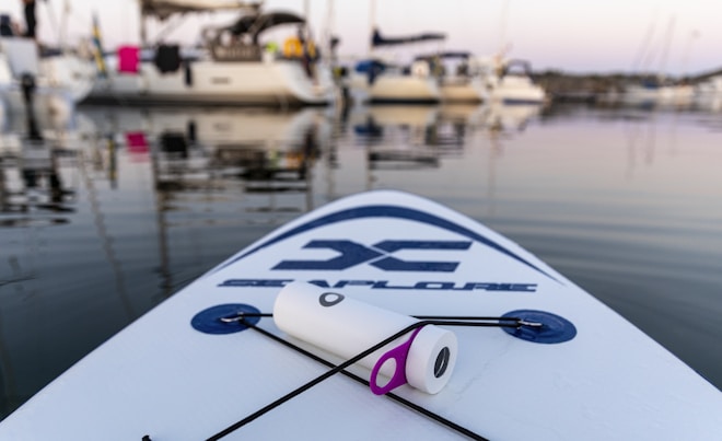 A stand-up paddleboard rests on calm waters in a marina, with several boats docked in the background. The paddleboard has a logo on it, and a cylindrical object with a purple ring is placed on it, secured by a rope. The sun is setting, casting a serene light over the scene.